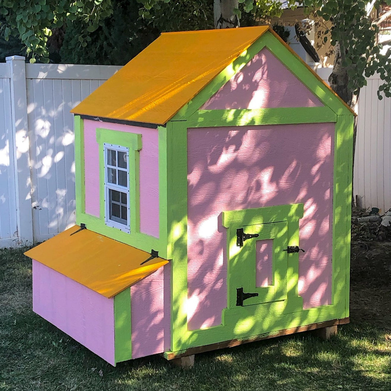 Colorful chicken coop with pink walls, green trim, and yellow roof in a backyard setting.